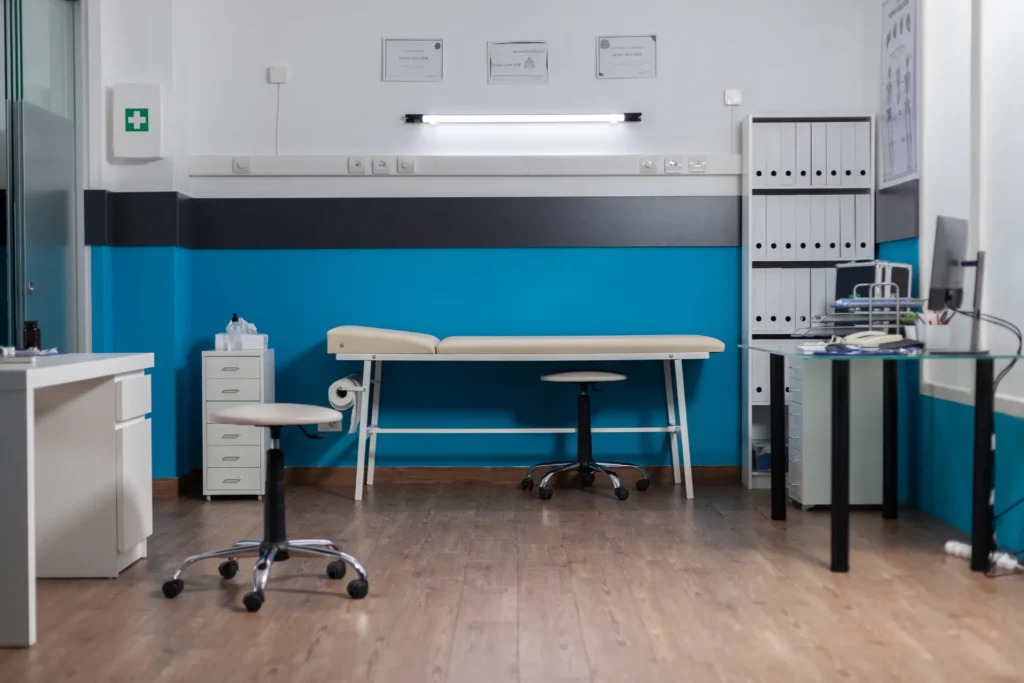 Salle de consultation médicale avec table d'examen, bureaux, fauteuil roulant et étagères pour documents et matériel médical.