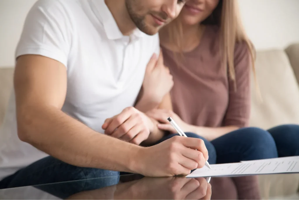 Un homme signe un contrat sur une table tandis qu'une femme lui pose tendrement la main sur l'épaule