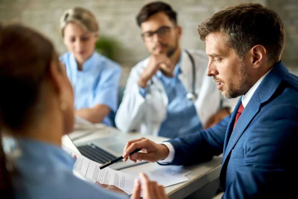 Un avocat en costume bleu pointe du doigt un document lors d'une réunion avec deux médecins et une infirmière.