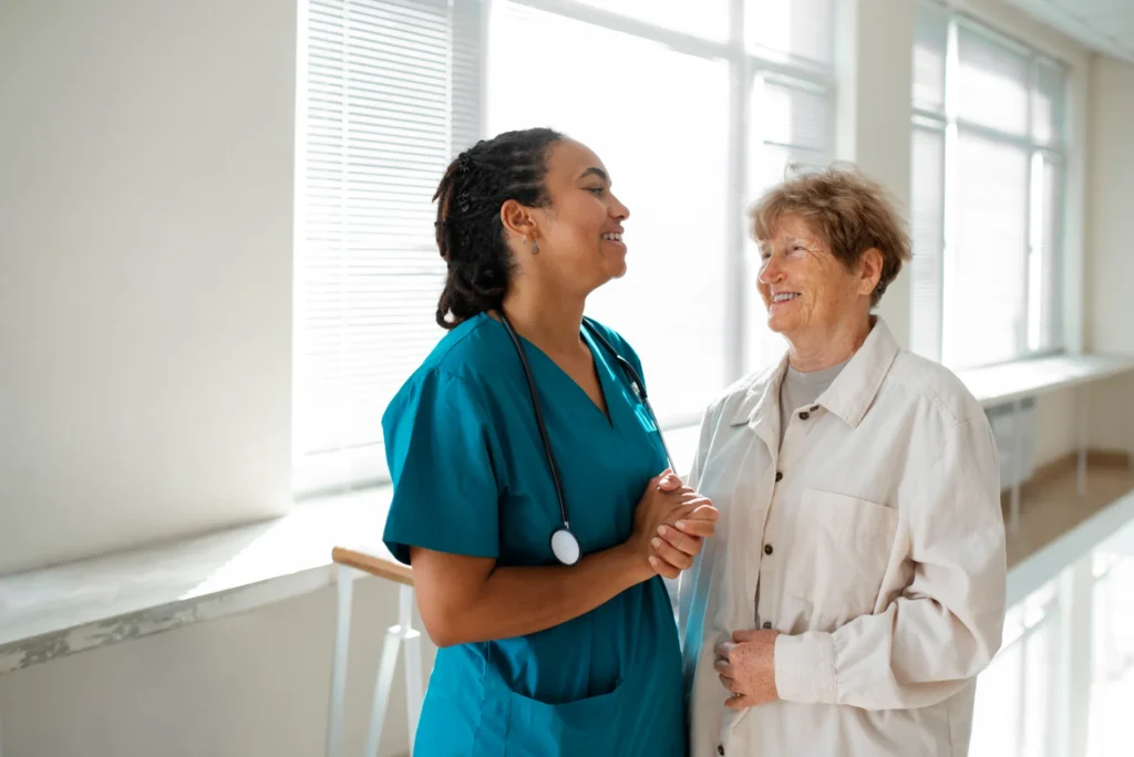 Uneaufnahme de deux femmes, une jeune soignante portant un uniforme bleu sarcelle et un stéthoscope, et une patiente plus âgée souriante, main dans la main, discutant dans un couloir lumineux de l'hôpital.