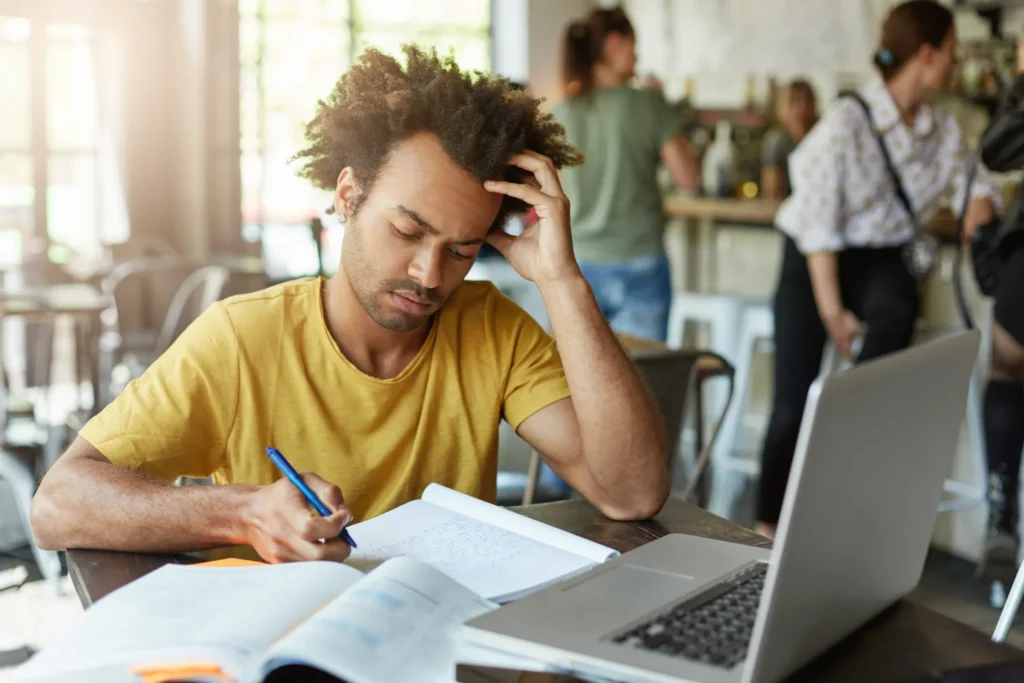 Un jeune homme aux cheveux bouclés étudie intensément dans un café avec un ordinateur portable et des livres ouverts devant lui.