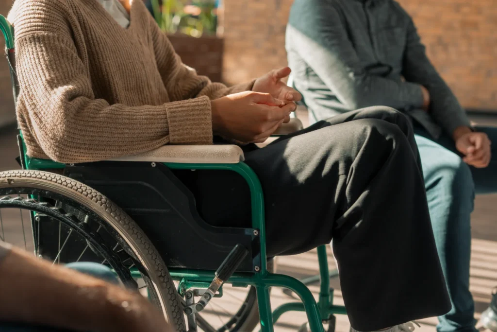 Gros plan sur une personne assise dans un fauteuil roulant, les mains jointes, alors qu'elle discute avec une autre personne assise à proximité.